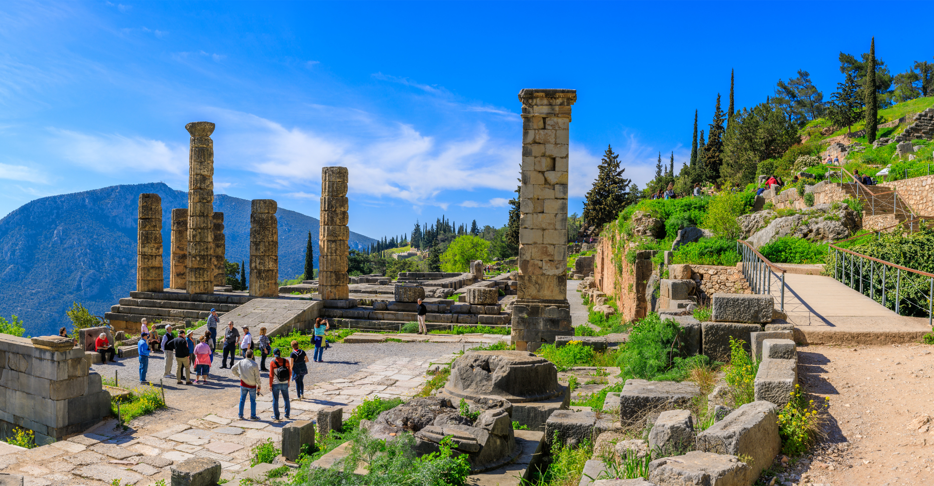 Tourists visiting ancient stone columns ruins at Delphi archaeological site under clear blue sky